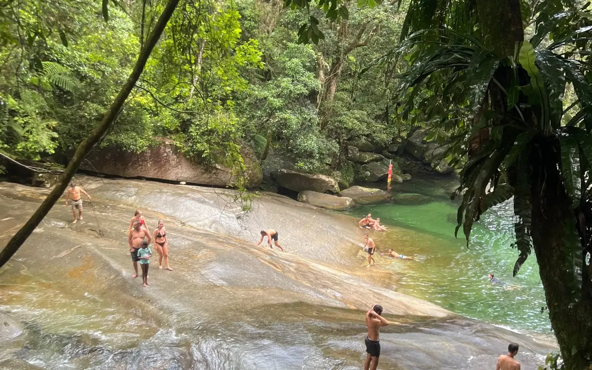 People standing and sitting on a wide smooth granite slab beside a green pool in rainforest