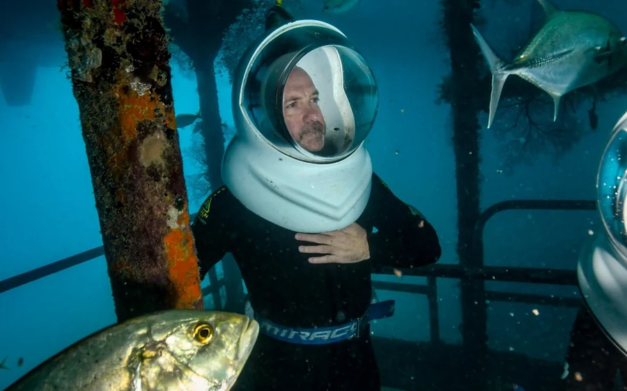 A diver in a clear helmet walking underwater among reef fish